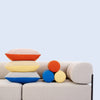 Frontaufnahme des nachhaltigen noah Sofa als Zweisitzer. Auf dem Sofa sind drei noah Kissen in Beigerot / Orange, Gelb / Gelb und Grau / Blau übereinander gestapelt. rechts daneben sind die dazugehörigen Nackenrollen zu sehen. 