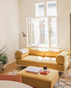 Nachhaltiges Zweisitzer noah Sofa in Senfgelb. Gestell in der Farbe weiß. Als Couchtisch wird ein großes Sitzmodul (95) ind der Farbe Senfgelb mit dem Gestell in Beigerot verwendet. Das Sofa steht in einem minimalistisch eingerichteten Wohnzimmer